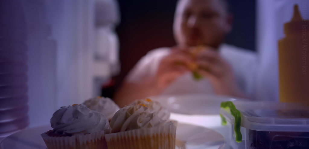 A photo of a man eating cupcakes from a fridge at night.