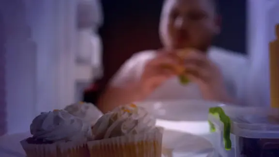 A photo of a man eating cupcakes from a fridge at night.